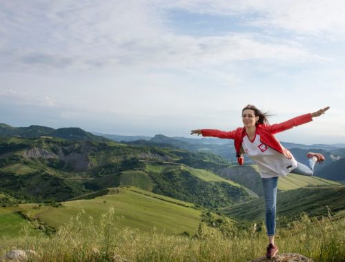 A woman joyfully balances on a rock amidst rolling green hills under a clear blue sky.